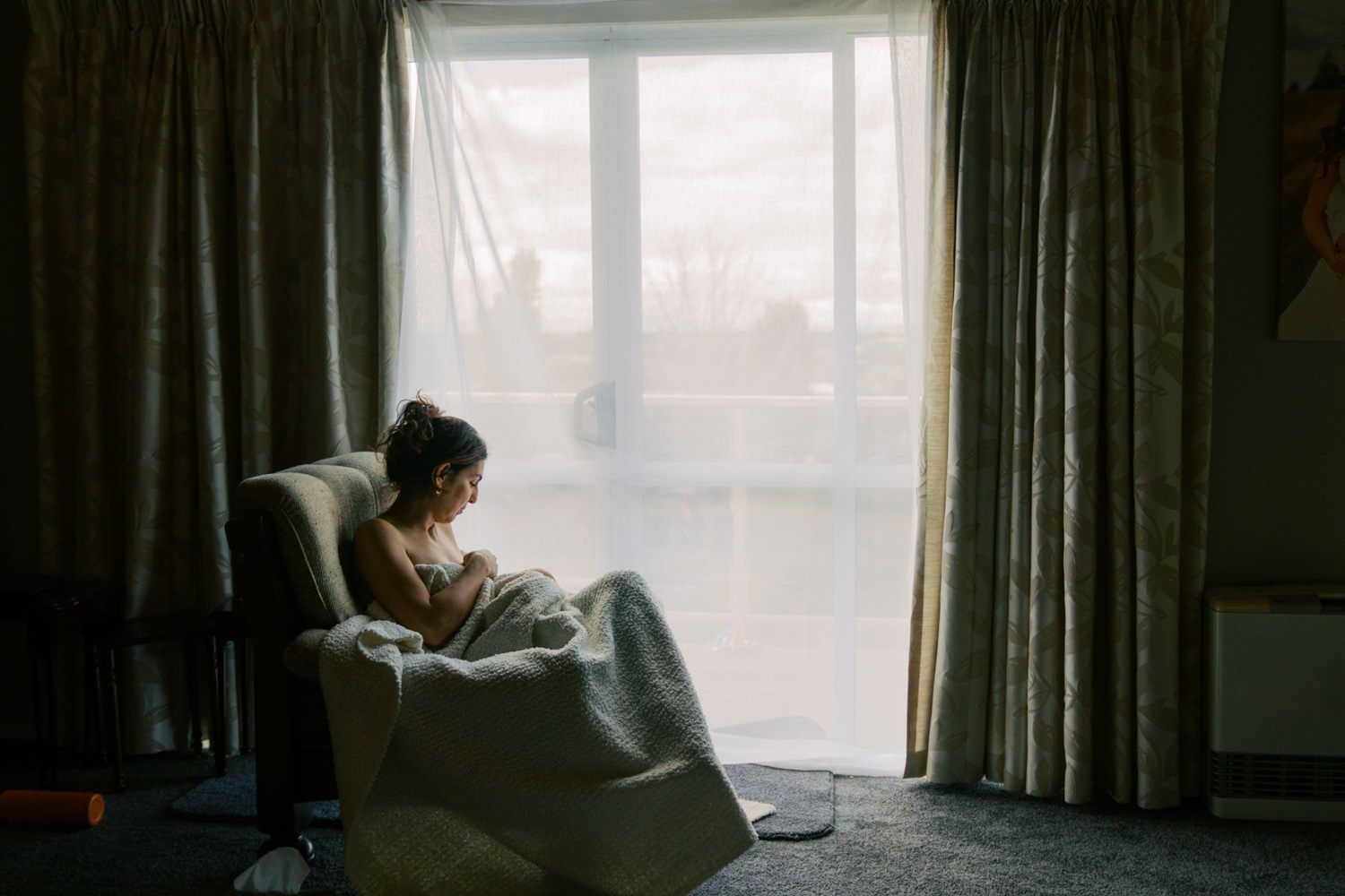 A young woman sits alone in an armchair by a bright window, wrapped in a blanket in a dimly lit room.