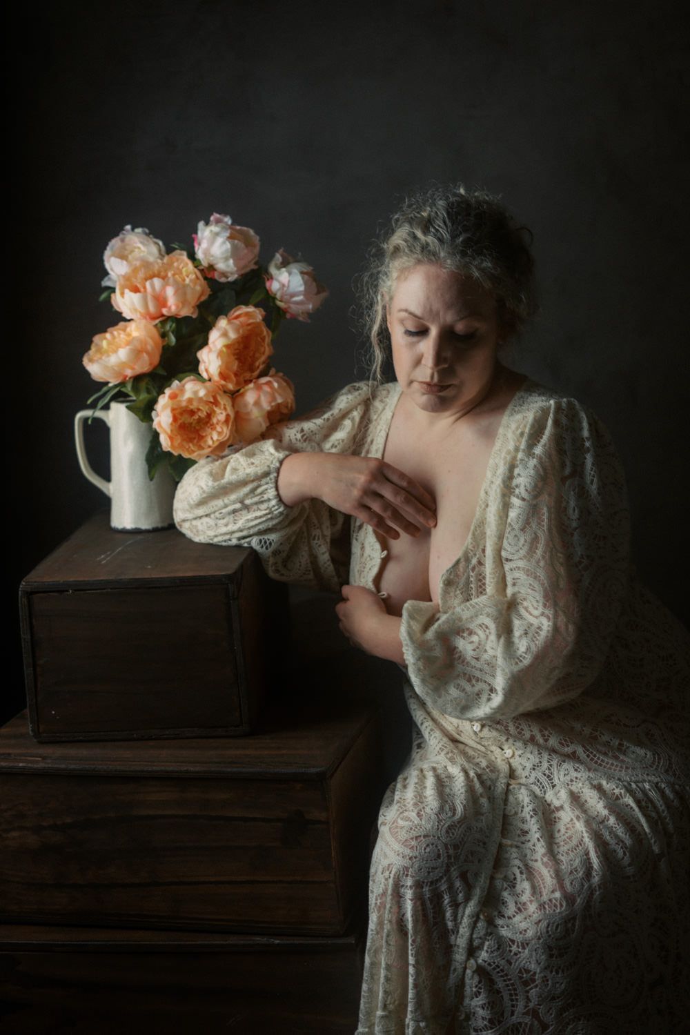 Woman in vintage lace gown posed beside pink peonies in white pitcher on antique wooden chest.