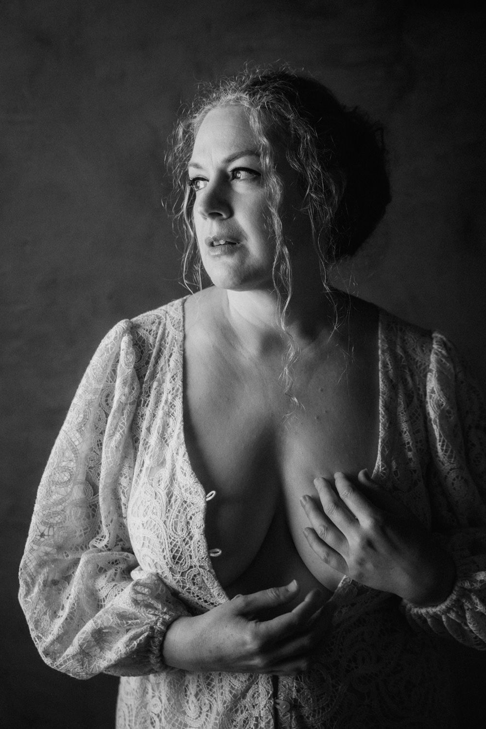 Black and white boudoir portrait of a woman in lace robe posing elegantly against a dark backdrop.