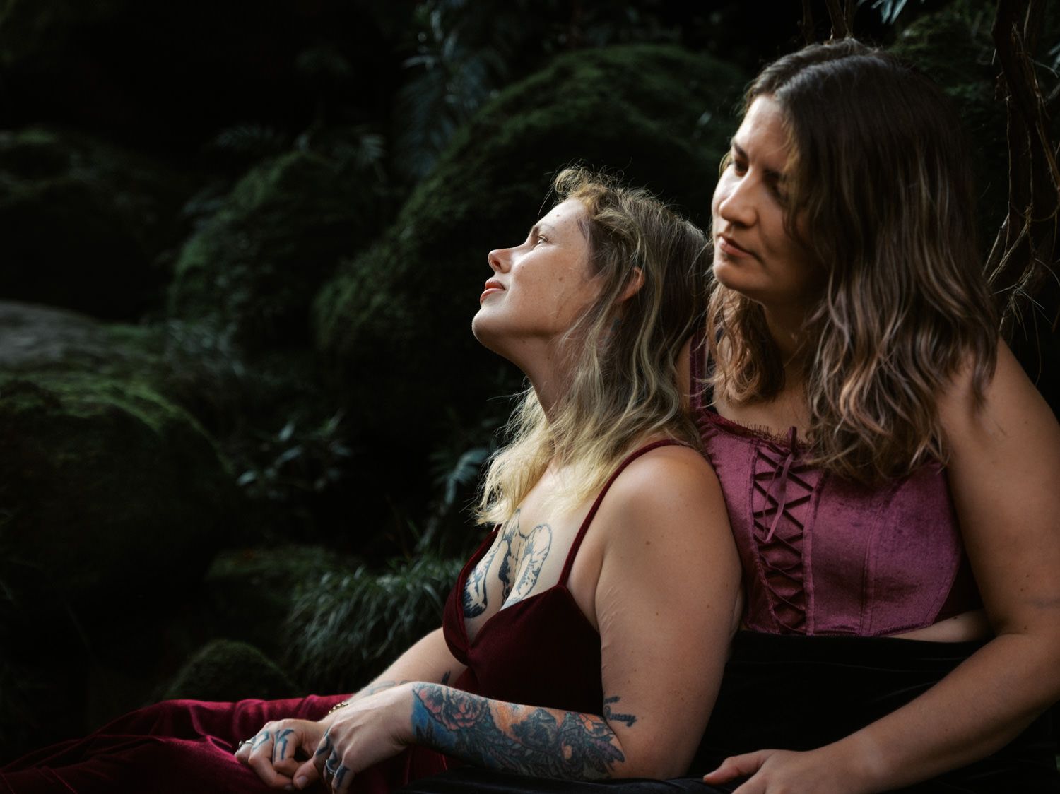 Two people in dark red velvet outfits pose romantically among moss-covered rocks in a lush forest setting.