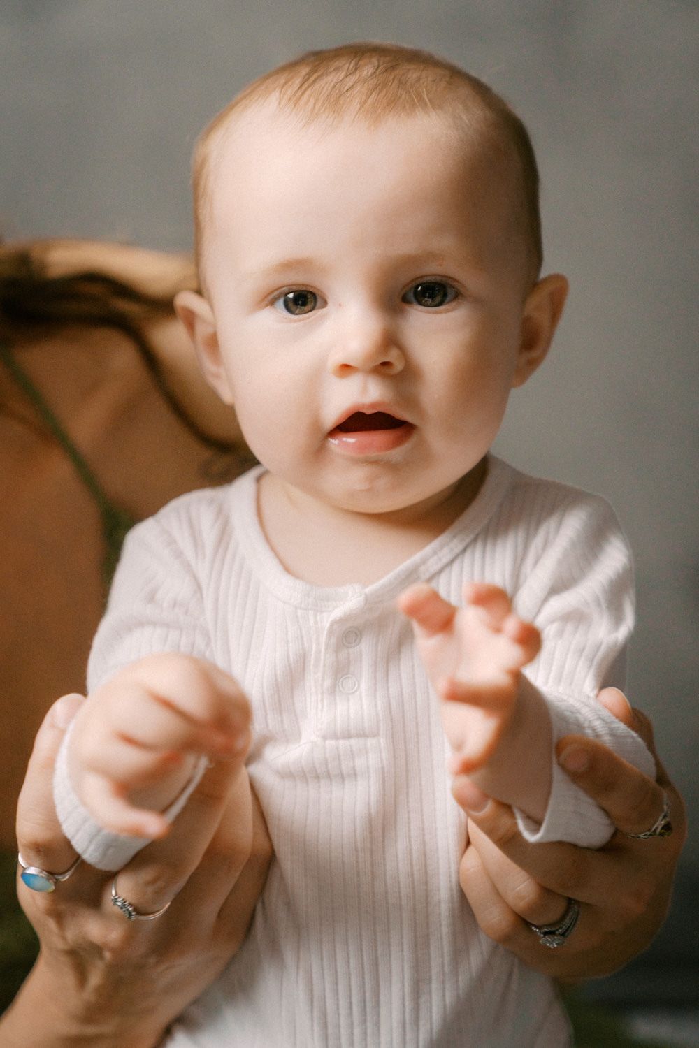 Adorable baby in white onesie reaching toward camera, supported by adult hands with silver rings.