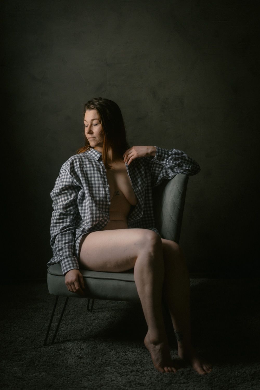 Woman in open plaid shirt seated in gray chair against dark studio background in artistic portrait.