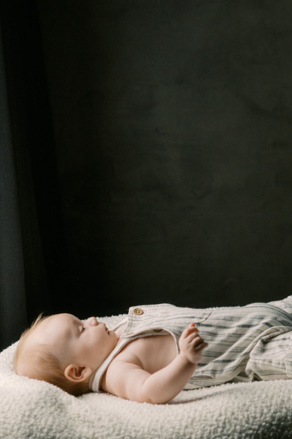 A peaceful baby in striped overalls lying on a fluffy white blanket against a dark background.