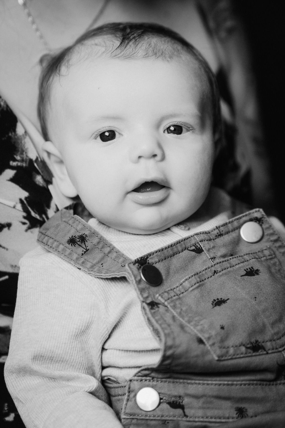 Black and white portrait of an adorable baby in denim overalls looking at the camera with wide curious eyes.