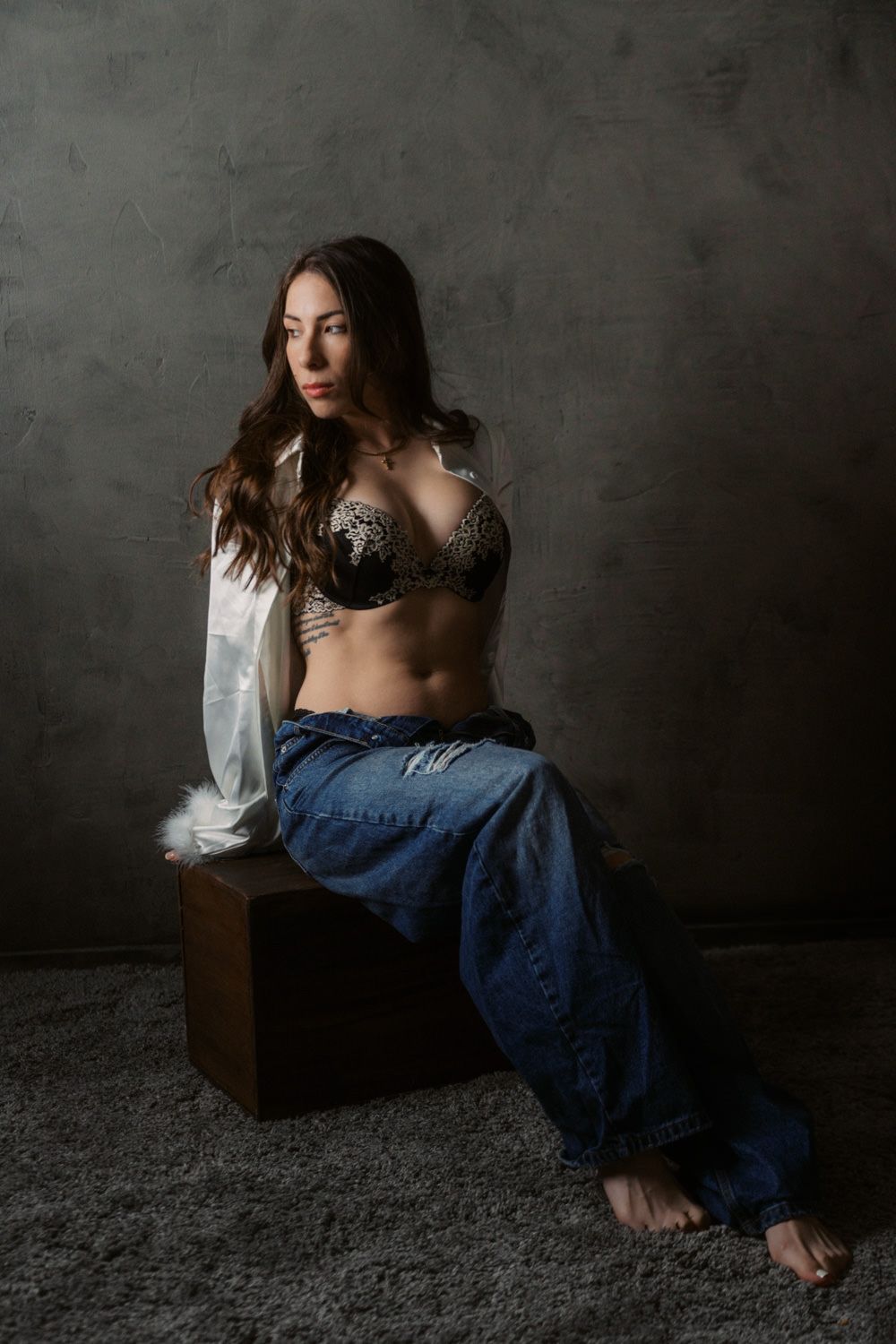 Woman in lace bra, open white shirt, and baggy jeans sits on wooden box in moody studio setting.