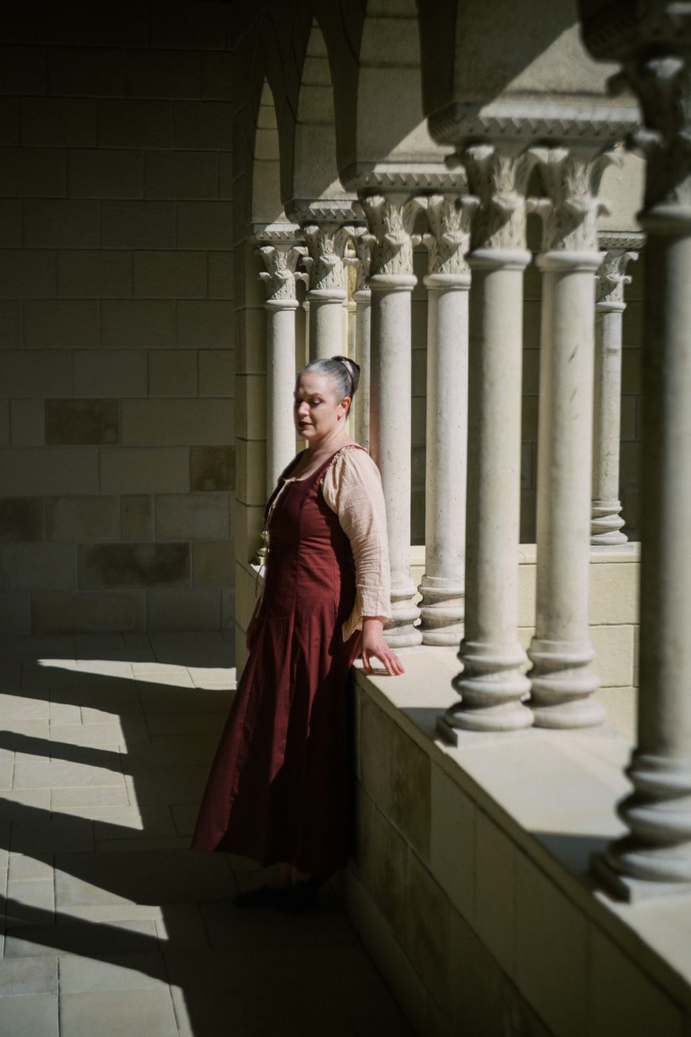 Woman in medieval dress standing among stone columns in a sunlit cloister arcade.