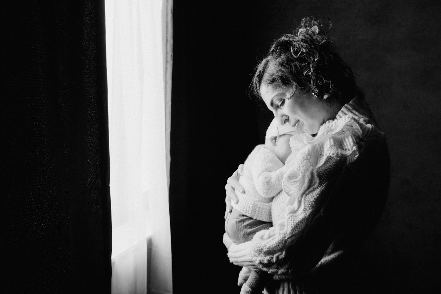 Black and white photo of a mother tenderly holding her newborn baby near a window in soft natural light.