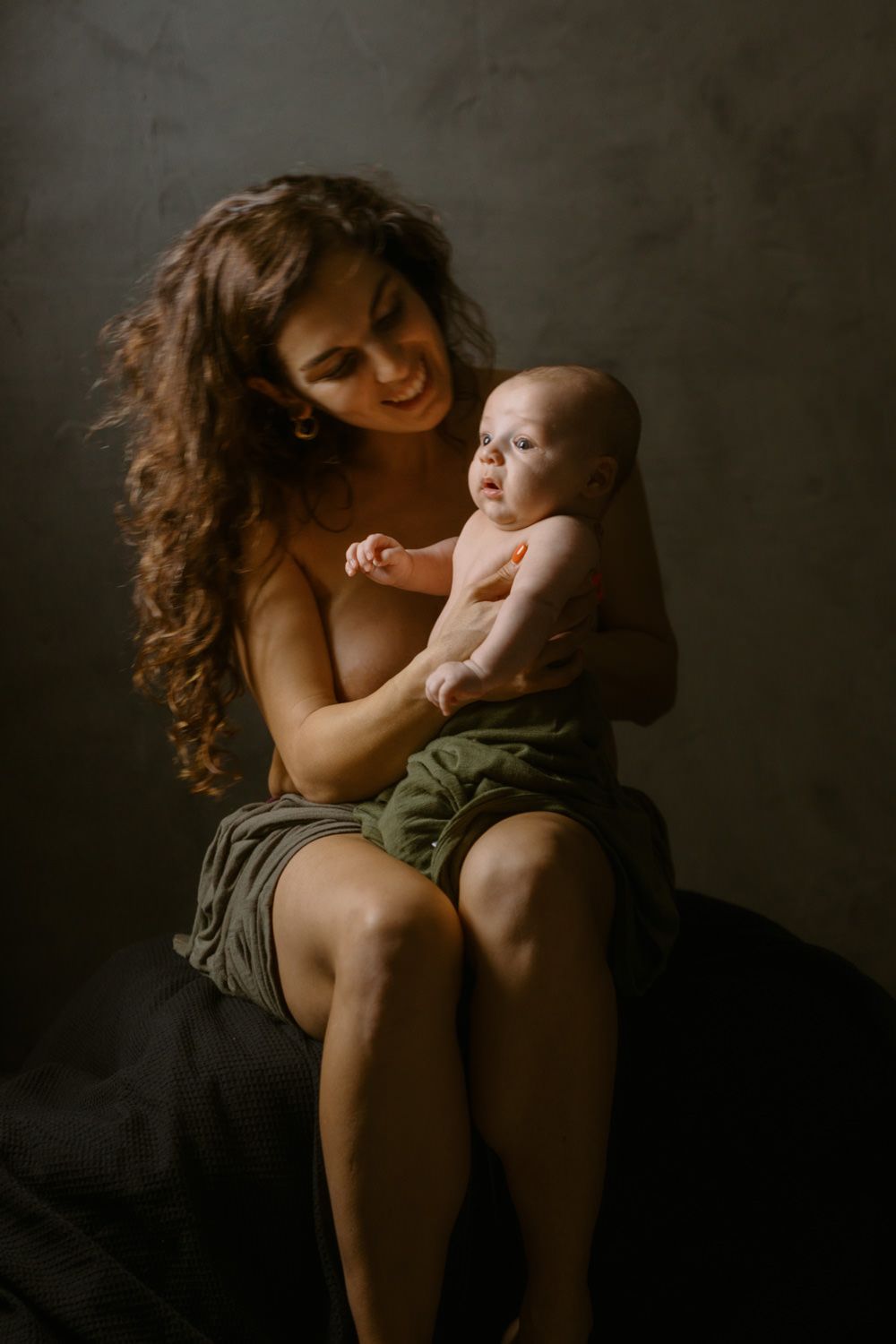 A smiling mother with curly hair holds her newborn baby while sitting, both wearing minimal clothing in a studio.