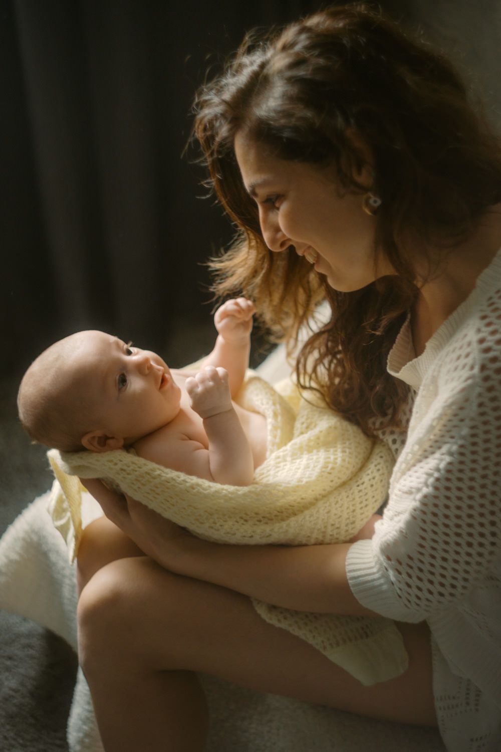 Young mother in white knit top holding newborn baby wrapped in yellow blanket in warm natural light.
