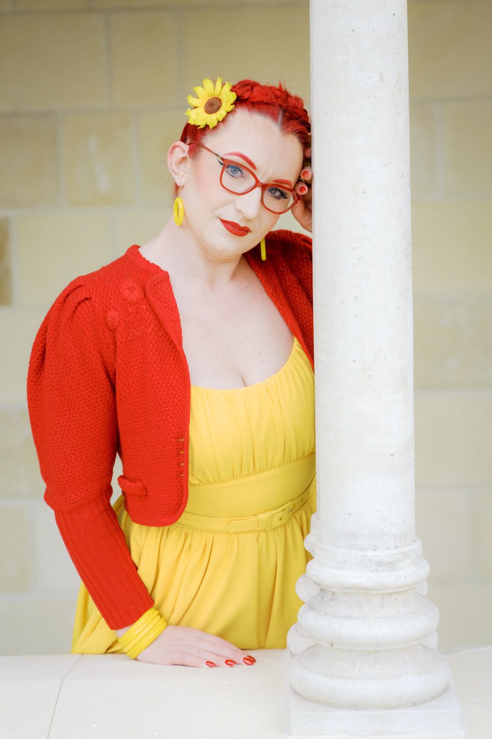 Woman in yellow dress and red cardigan with matching glasses and sunflower hair accessory leaning against white column.