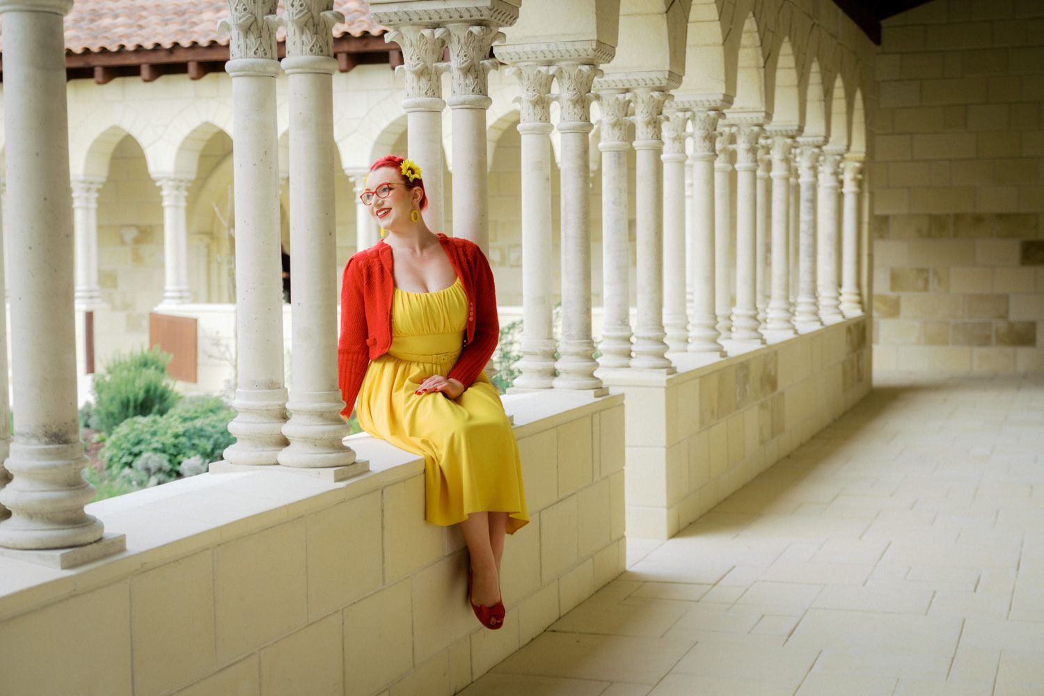 Woman in yellow dress and red cardigan sitting on white stone balustrade in elegant columned arcade.
