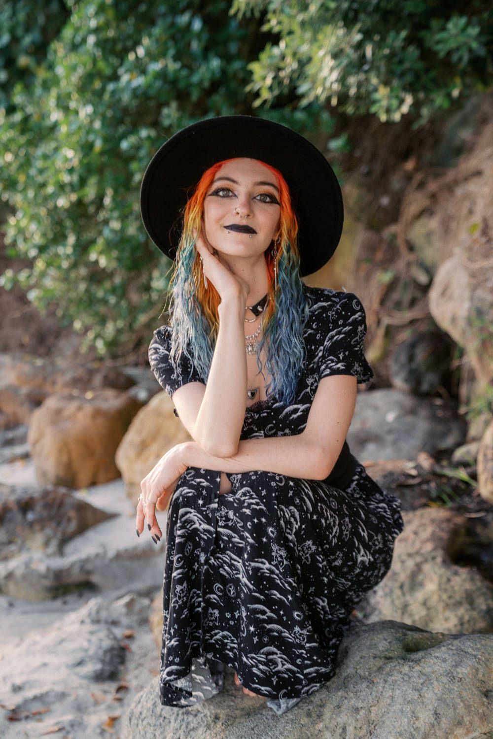 Woman with colorful red and blue hair, black hat, dark lipstick, and floral dress sitting on rocks outdoors.