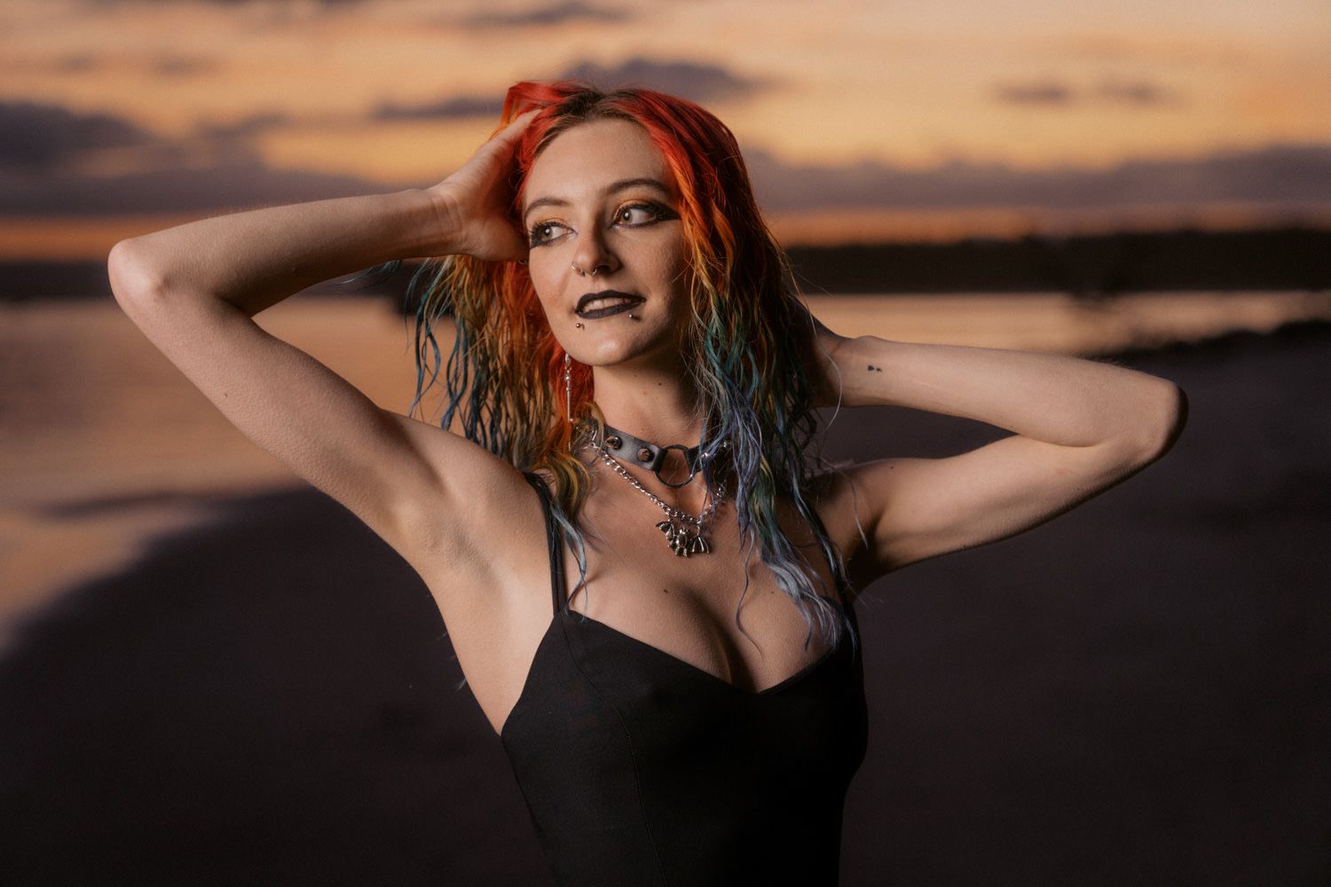 Woman with colorful red and blue hair poses in black dress at sunset beach, wearing statement necklace.