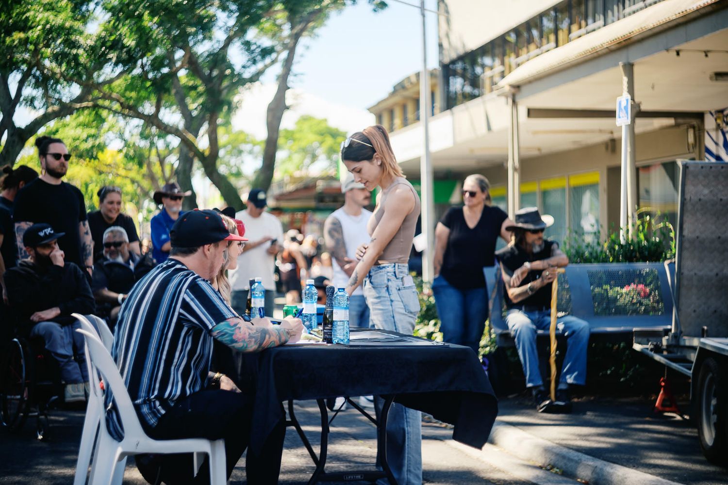 People gathered outdoors at a street event, with a woman standing at a table while others sit and socialize nearby.