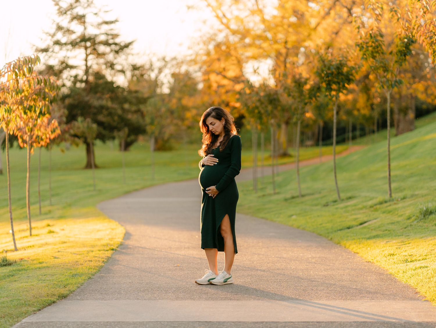 Pregnant woman in black dress cradling baby bump on a sunlit park path surrounded by autumn trees.