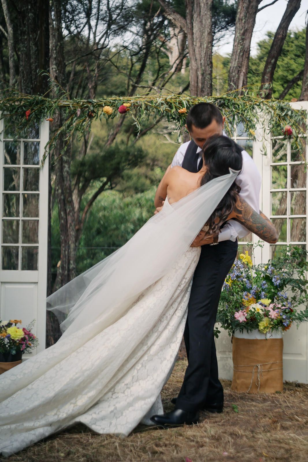 Bride and groom sharing first kiss at outdoor wedding ceremony with floral arch and white doors.