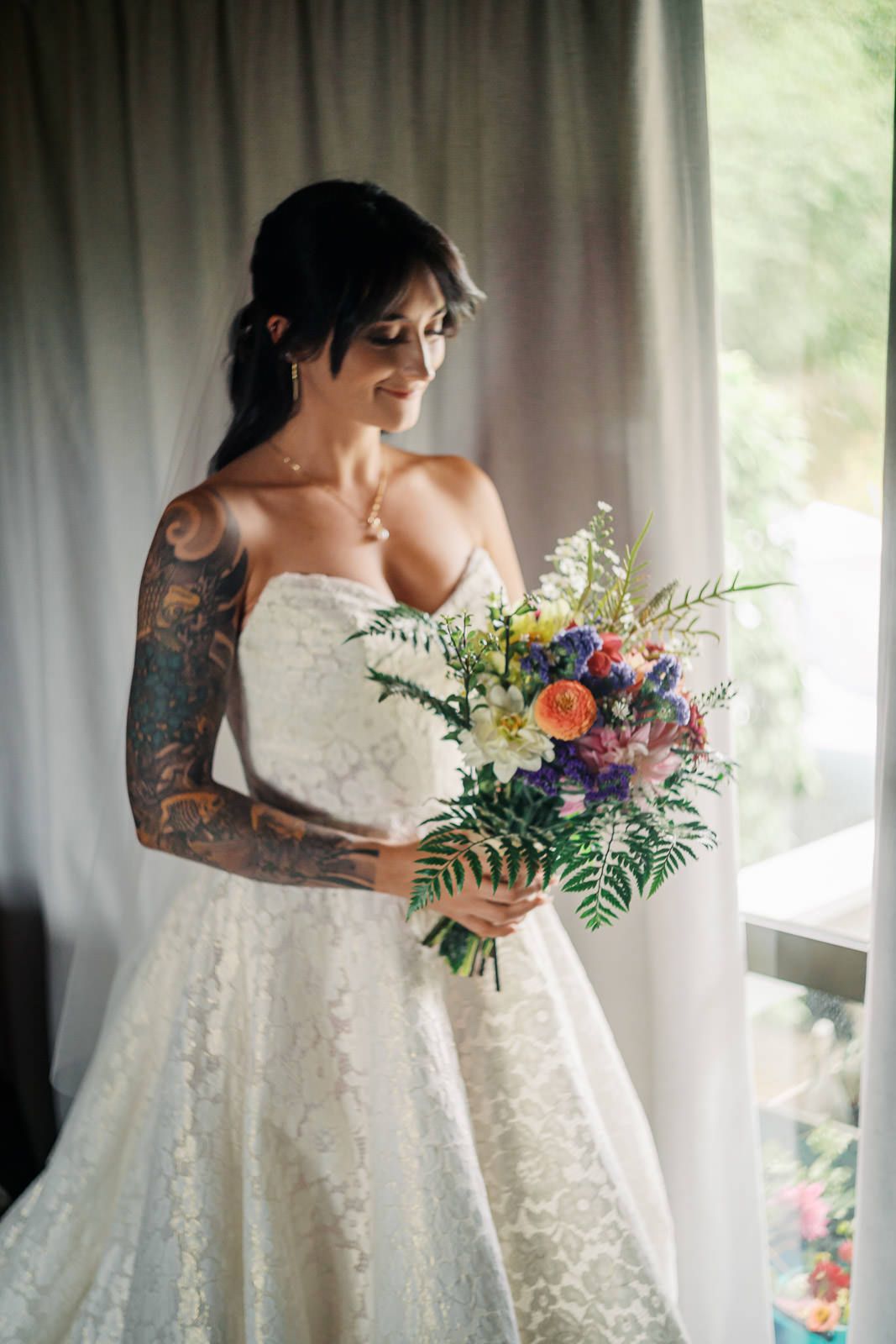 Tattooed bride in strapless lace wedding gown holds colorful bouquet of wildflowers, smiling softly by a sunlit window.
