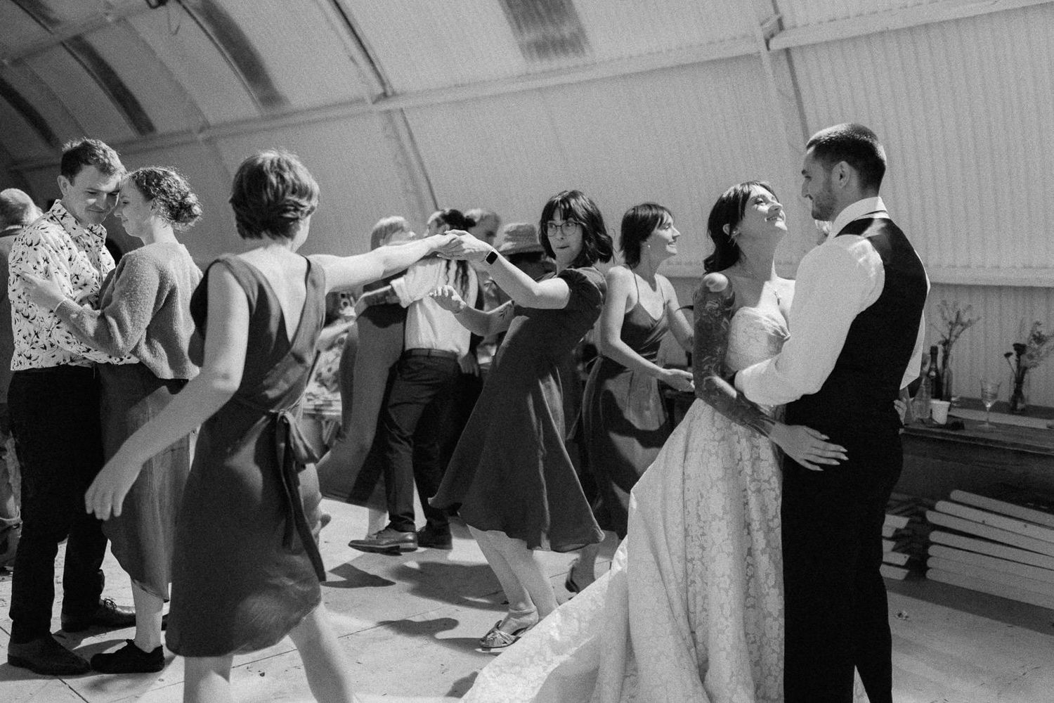 Black and white photo of wedding guests dancing joyfully at a reception, with bride and groom sharing a romantic moment.