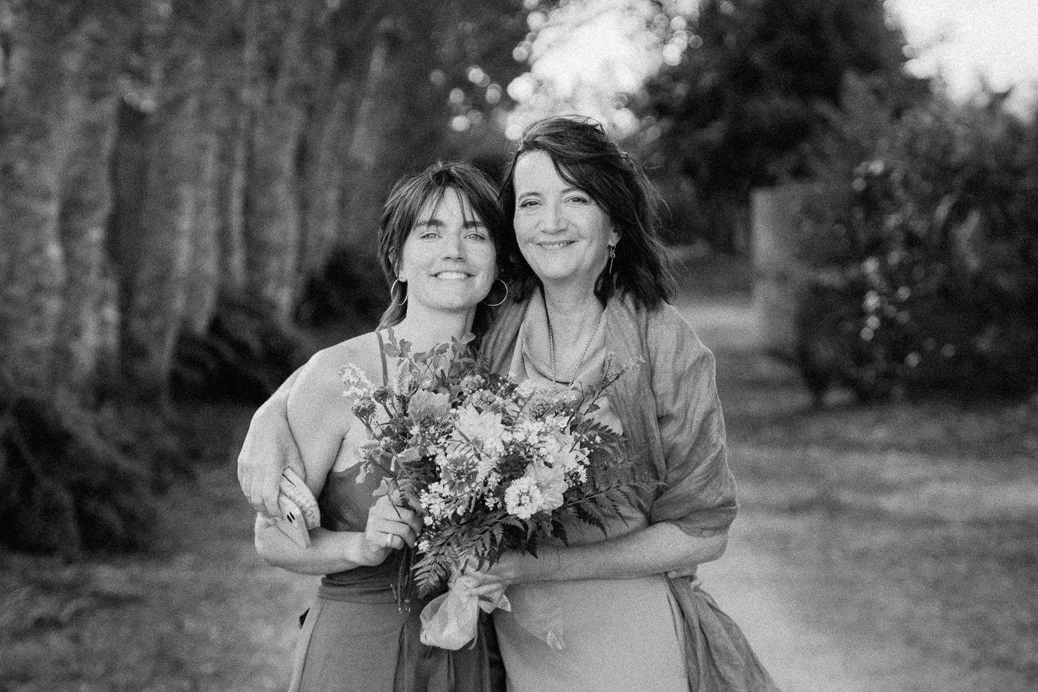 Two women smiling and posing together outdoors, one holding a bouquet of flowers, in a black and white photo.