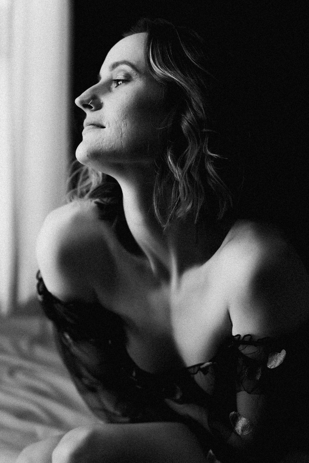 Black and white boudoir portrait of a woman in lace lingerie gazing toward window light.
