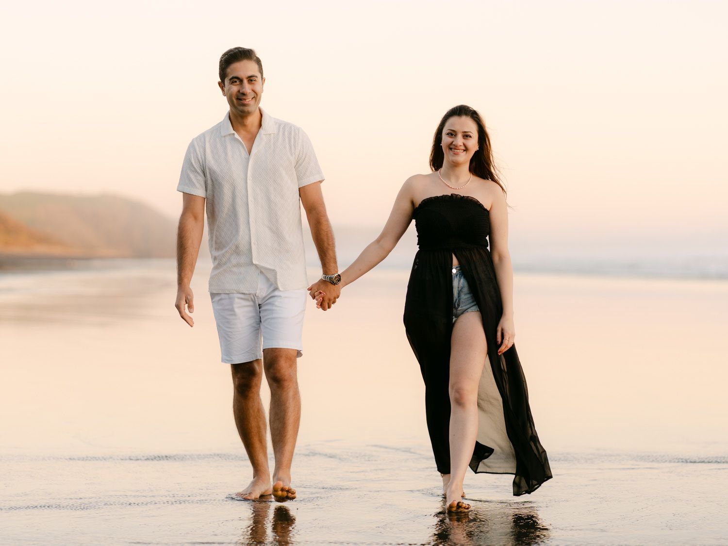 Couple holding hands walking barefoot on a beach at sunset, man in white shirt and shorts, woman in black strapless dress.