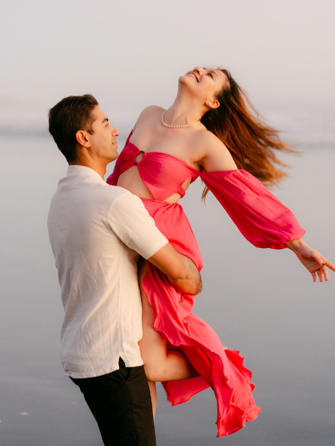 Man in white shirt lifts woman in pink flowing dress near calm water at sunset.