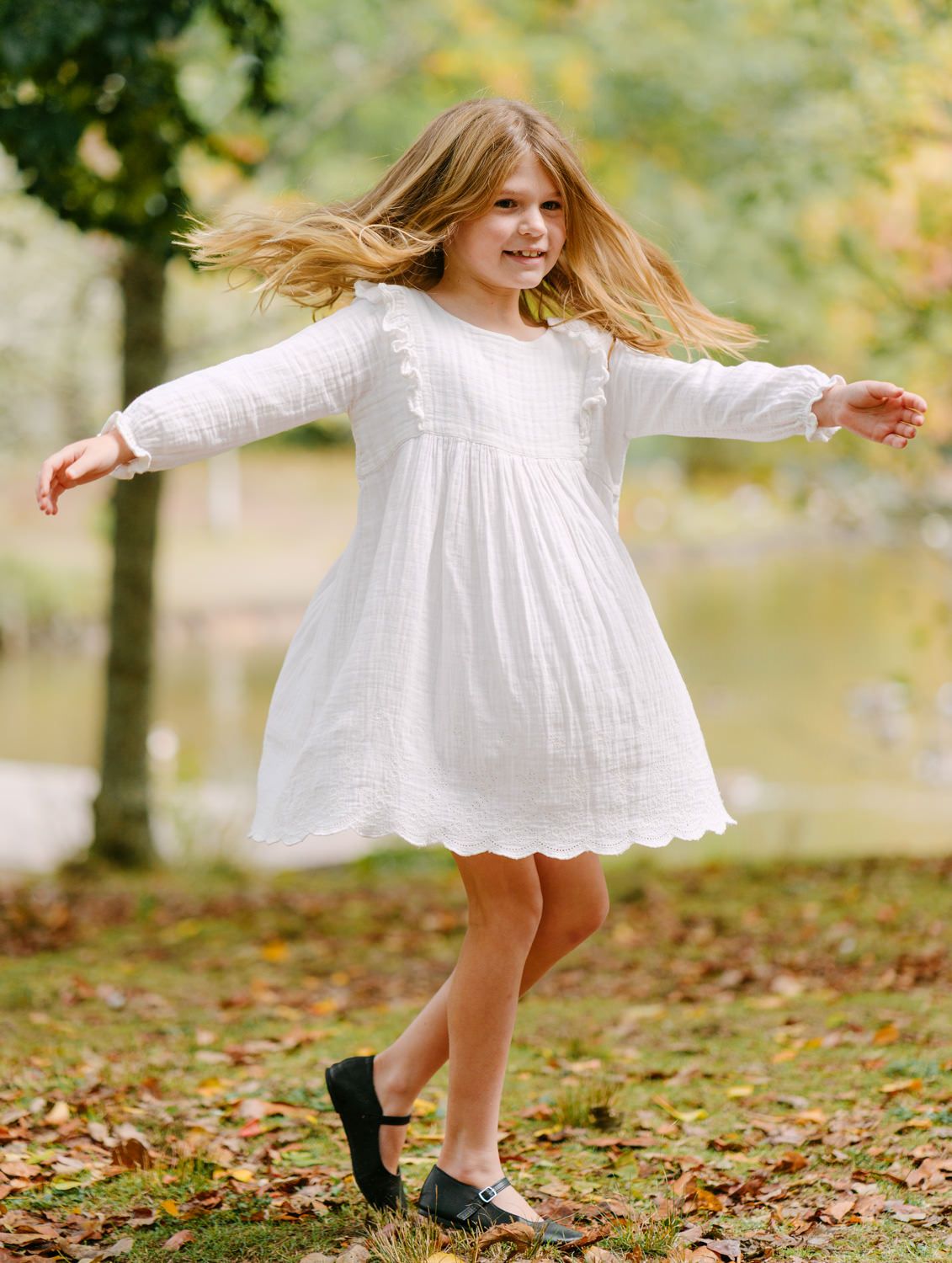 Young girl twirling in white long-sleeve scalloped dress with ruffles, playing outdoors in autumn park.