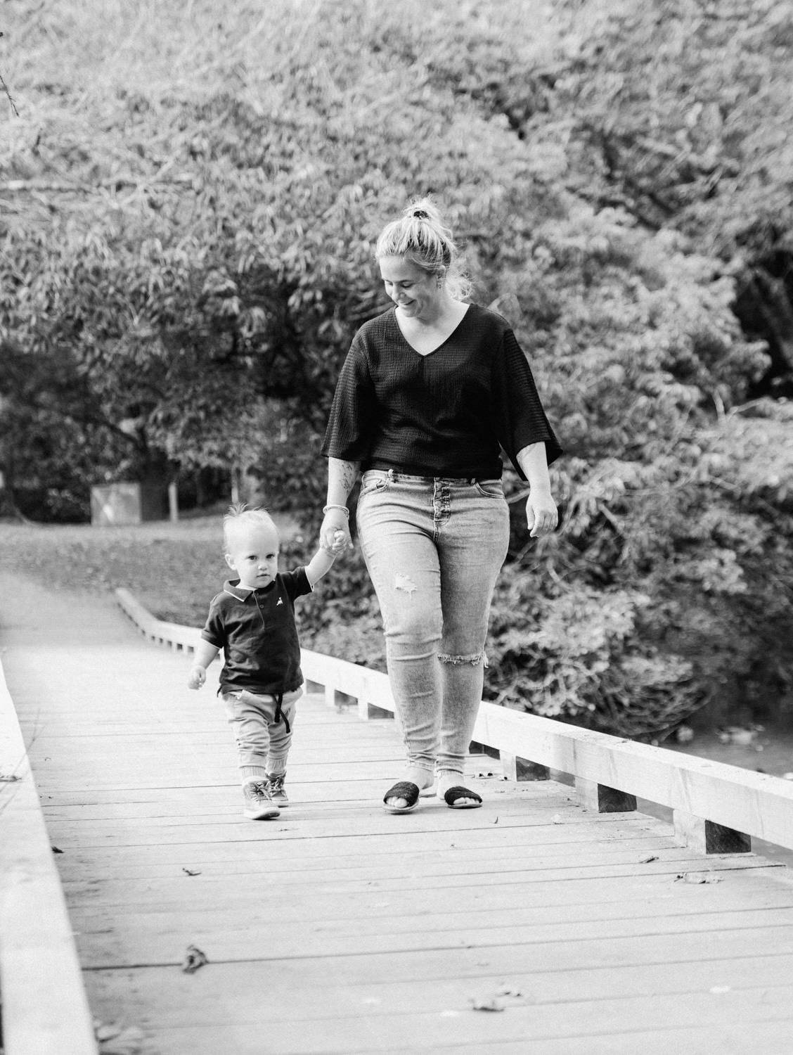 Black and white photo of a mother and toddler son walking hand in hand on a wooden bridge surrounded by trees.