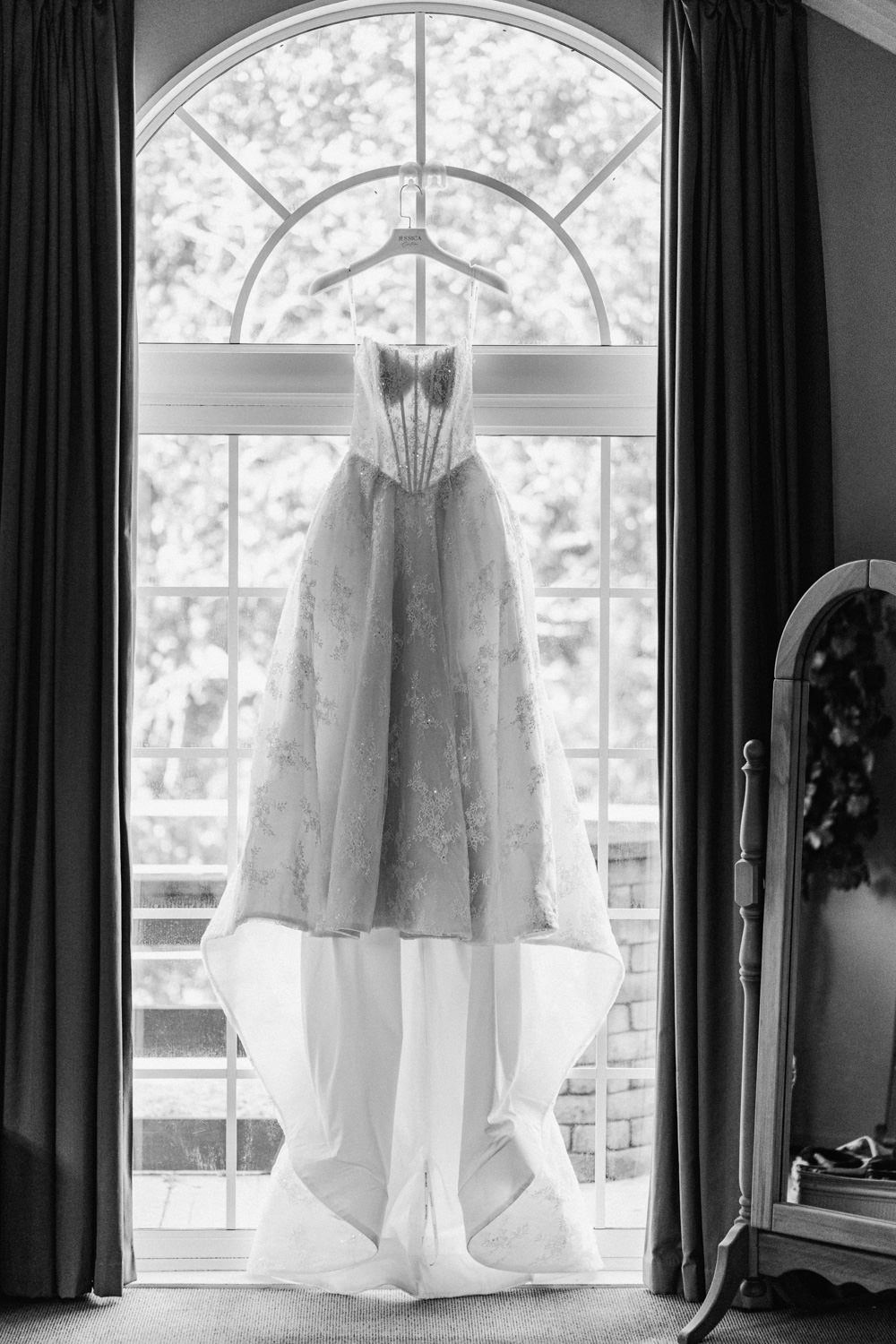 Black and white photo of an elegant wedding gown hanging in an arched window with flowing skirt and lace bodice details.
