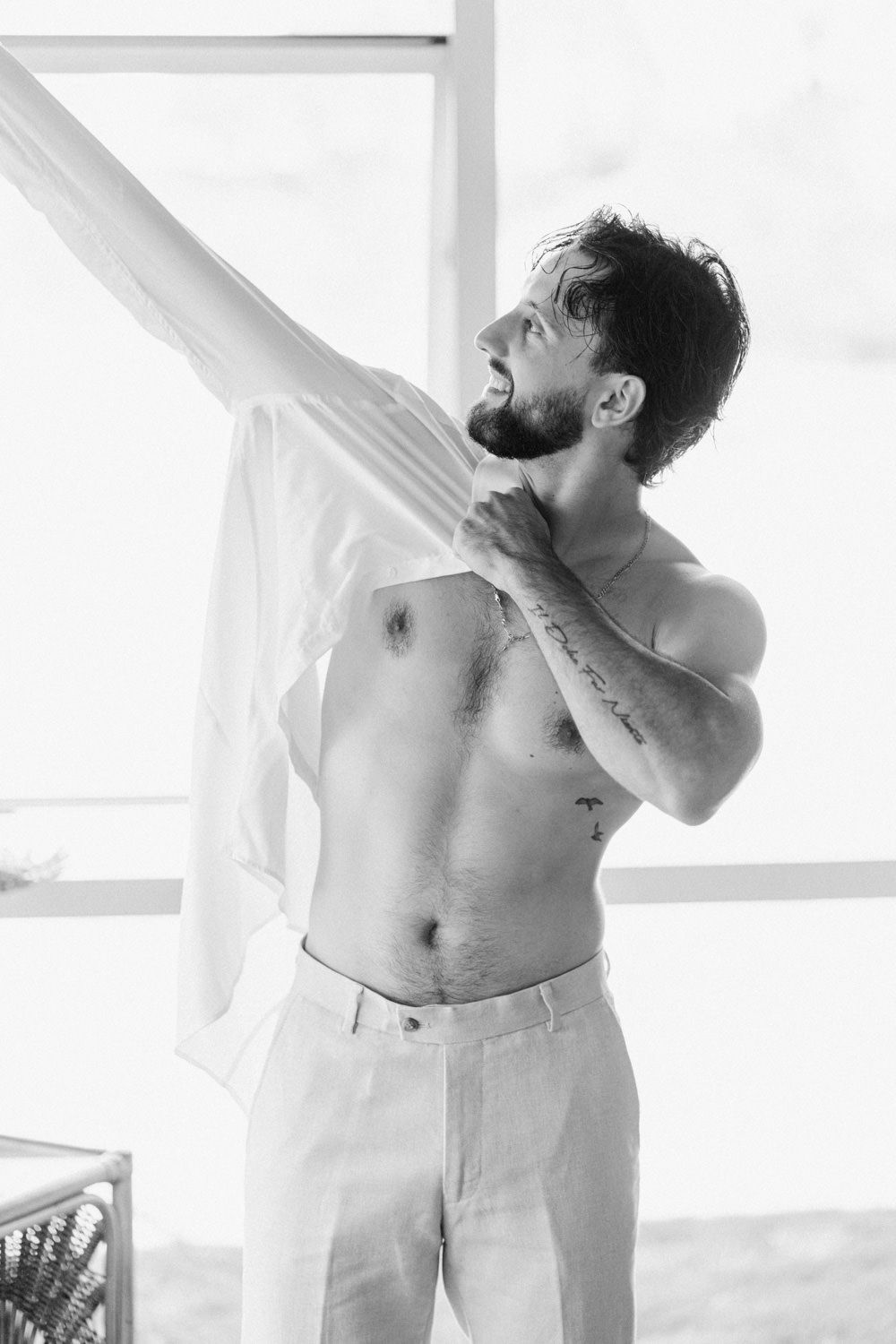 Shirtless bearded man holding white curtain in black and white photo, wearing light trousers with chest tattoo visible.