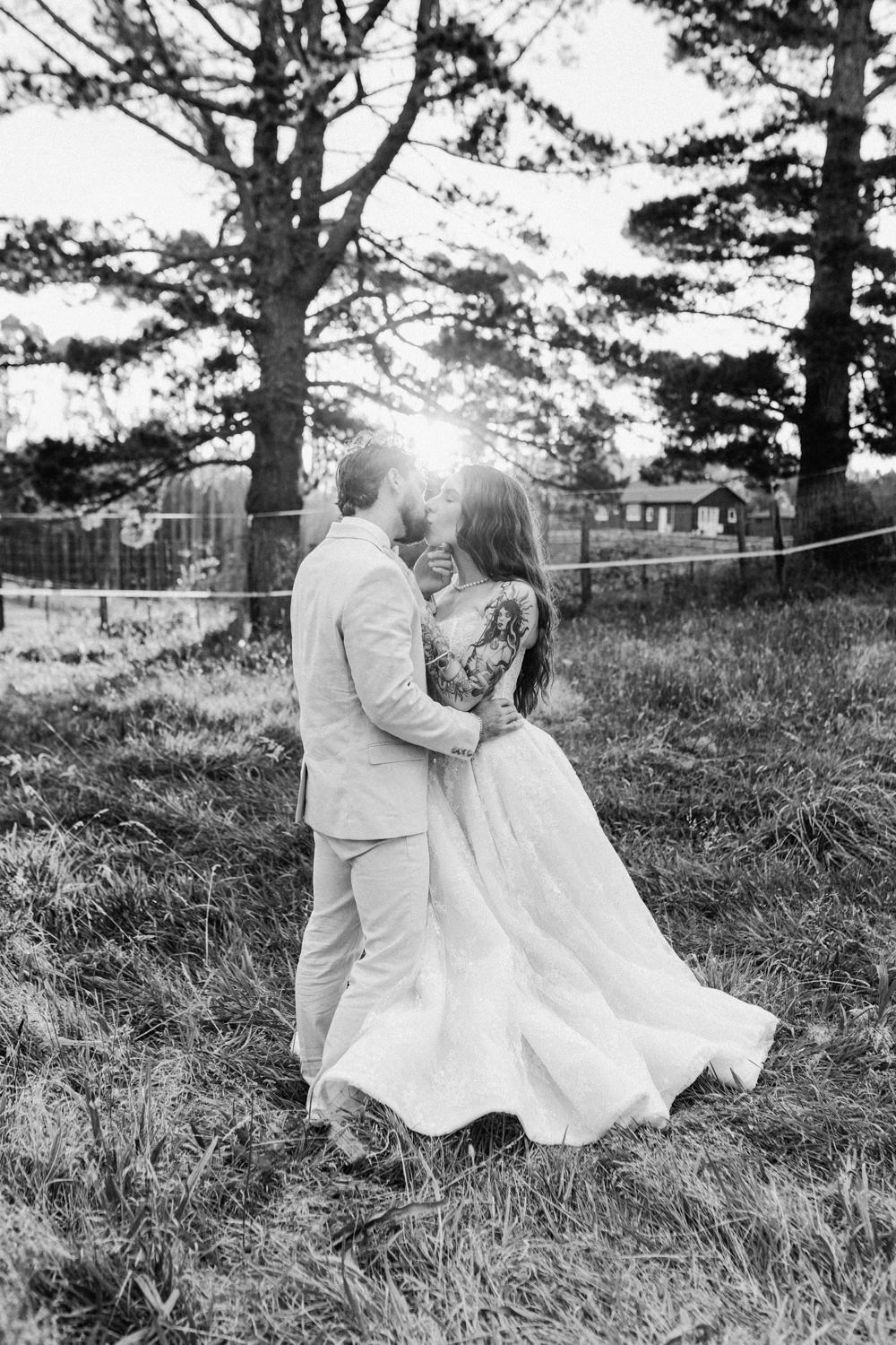 Black and white photo of a couple sharing a romantic kiss outdoors in a field, bride in flowing gown, sun shining through trees.