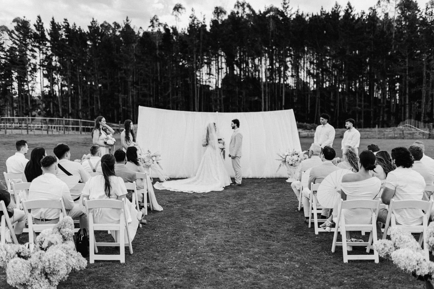 Black and white outdoor wedding ceremony with bride and groom at altar, guests seated in white chairs, pine trees backdrop.