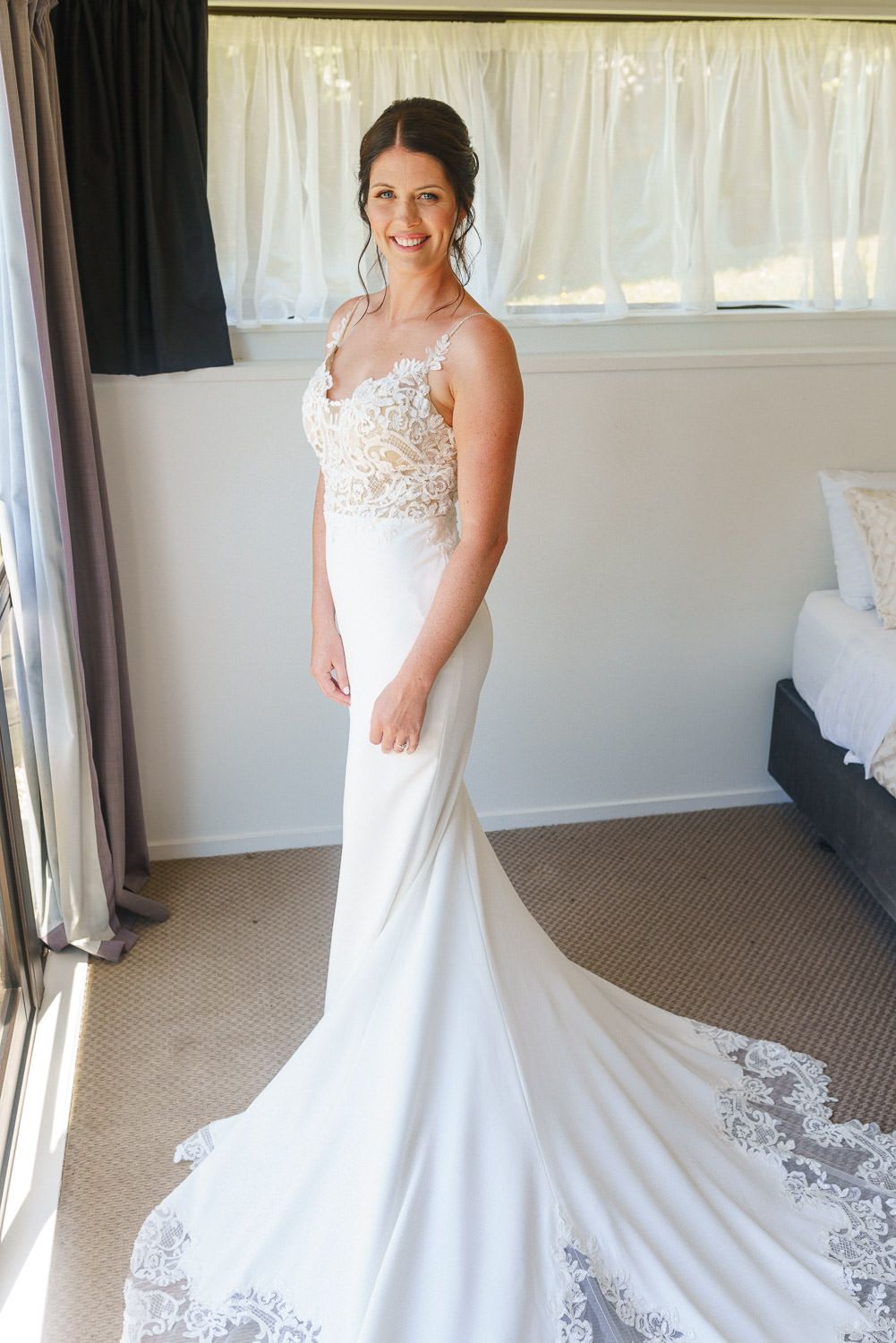 Smiling bride in elegant white lace mermaid wedding gown with train, posing in a bright room.