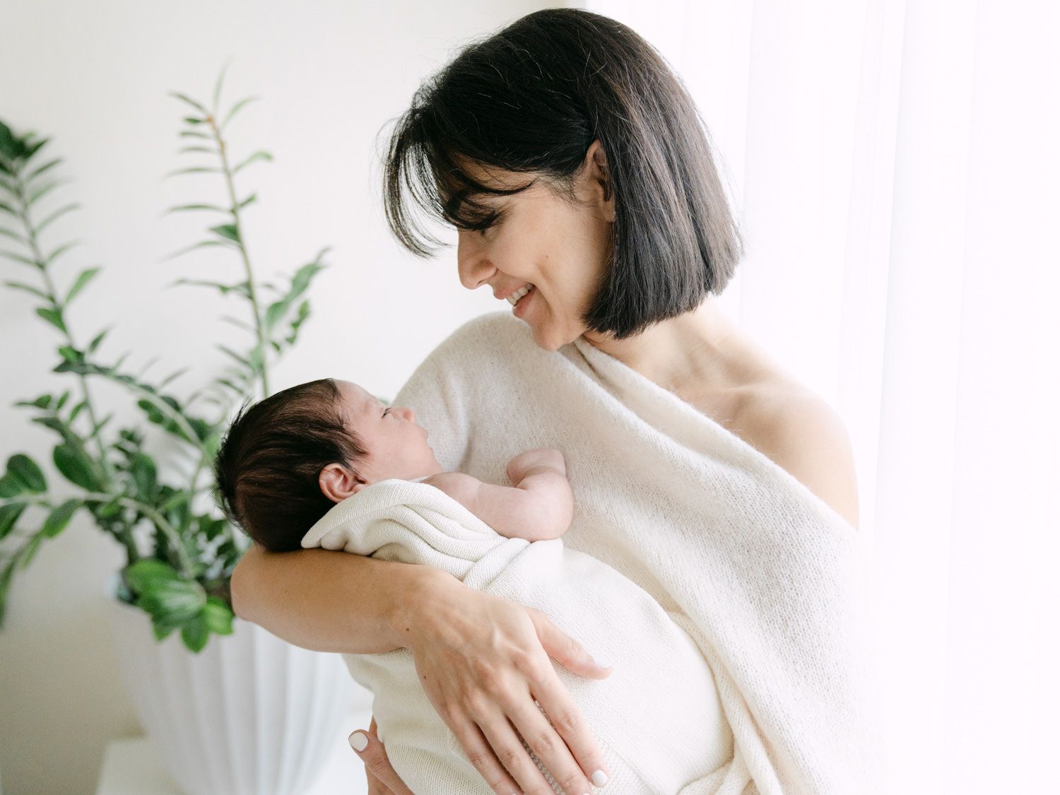Smiling mother gazing lovingly at her swaddled newborn baby in bright room.