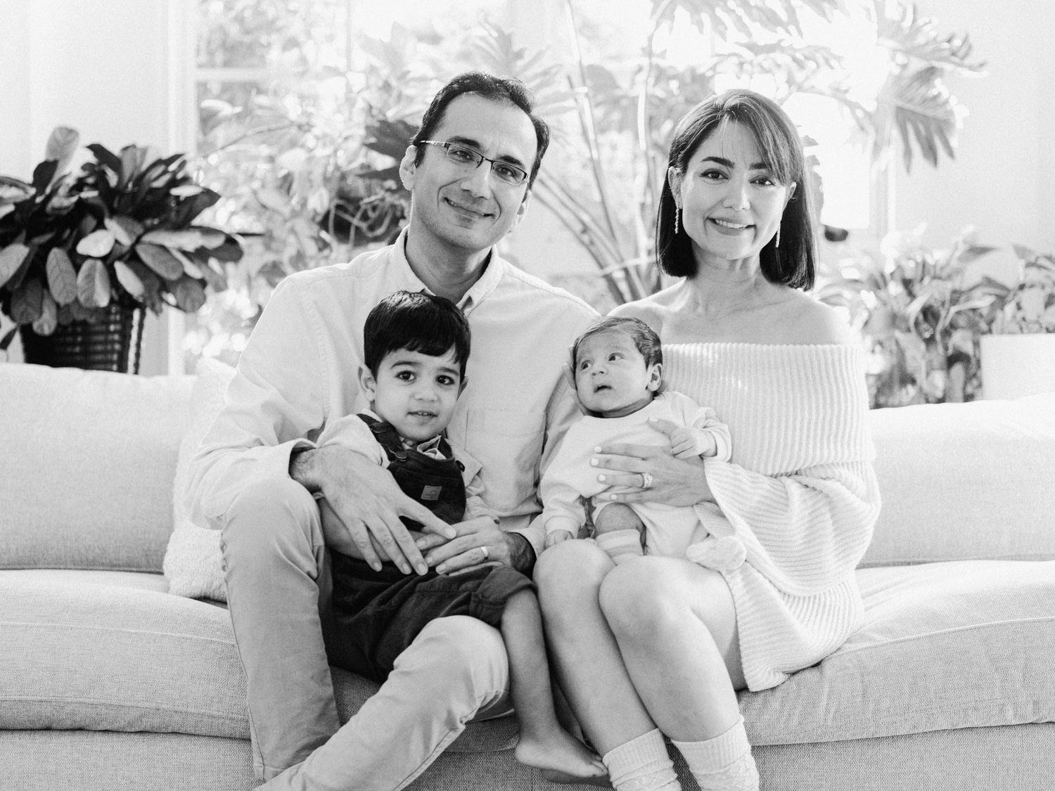 A family of four poses together in a black and white portrait on a white sofa indoors.