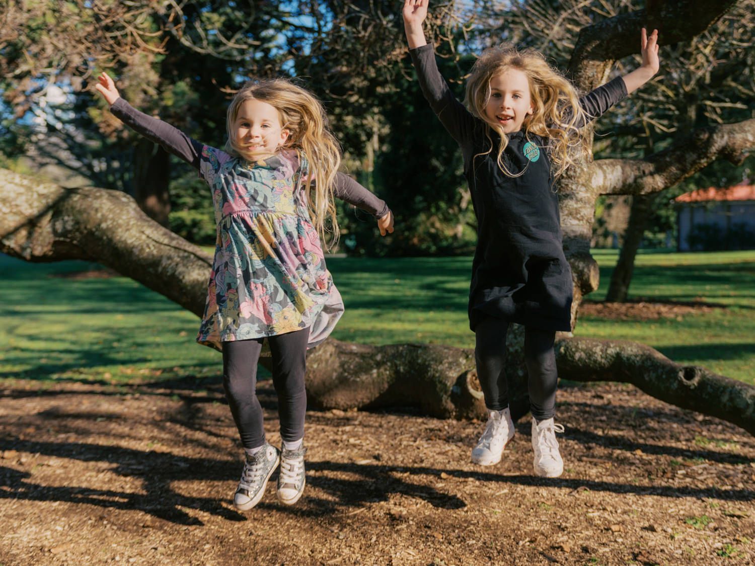 Two young girls jump joyfully in the air with arms raised in a colorful outdoor park setting.