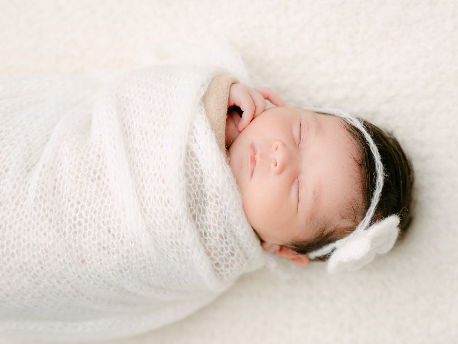 Peaceful newborn baby swaddled in white knit wrap with floral headband sleeping on soft white blanket.