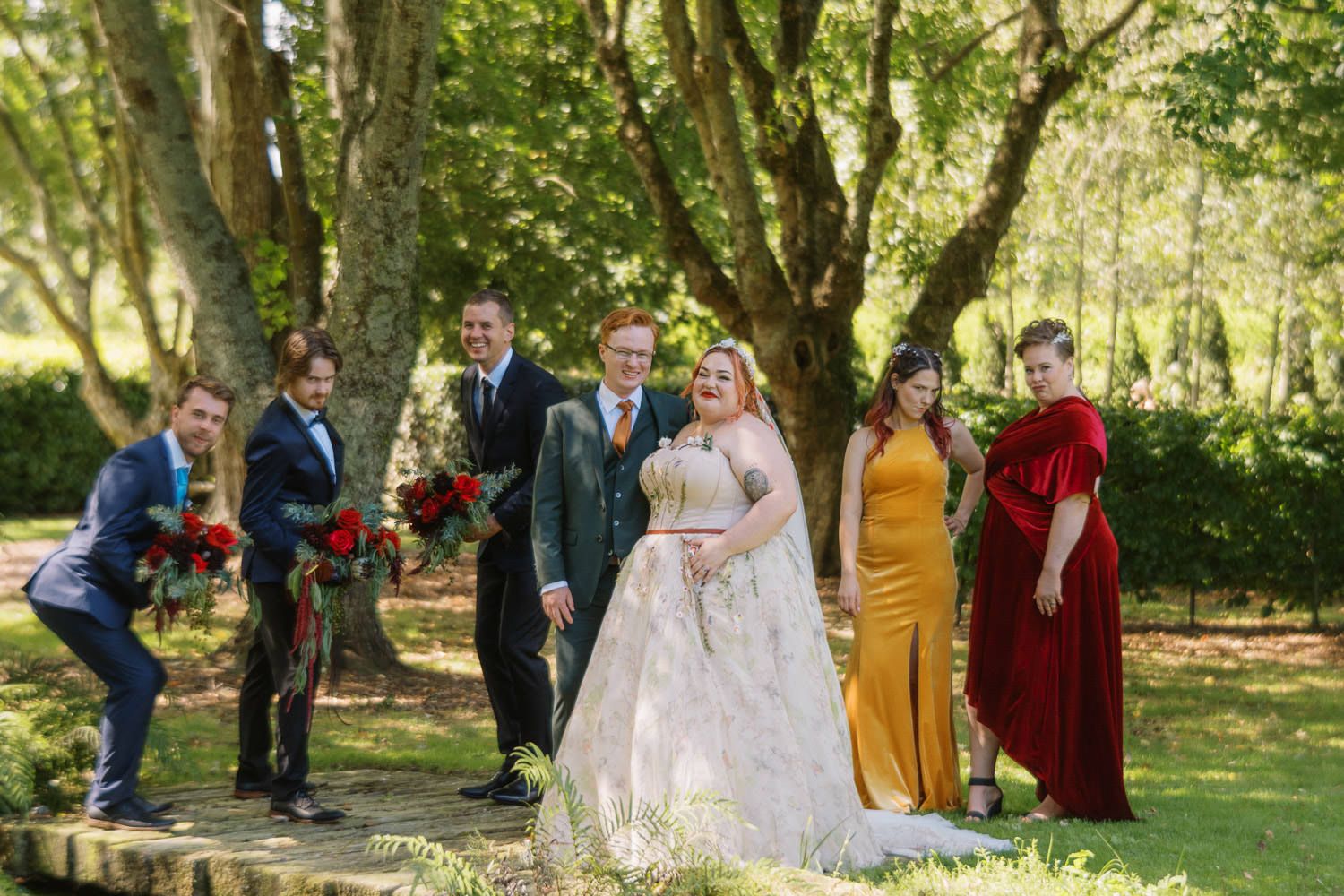 Wedding party poses outdoors among lush green trees, bride in white gown with groomsmen and bridesmaids in colorful dresses.