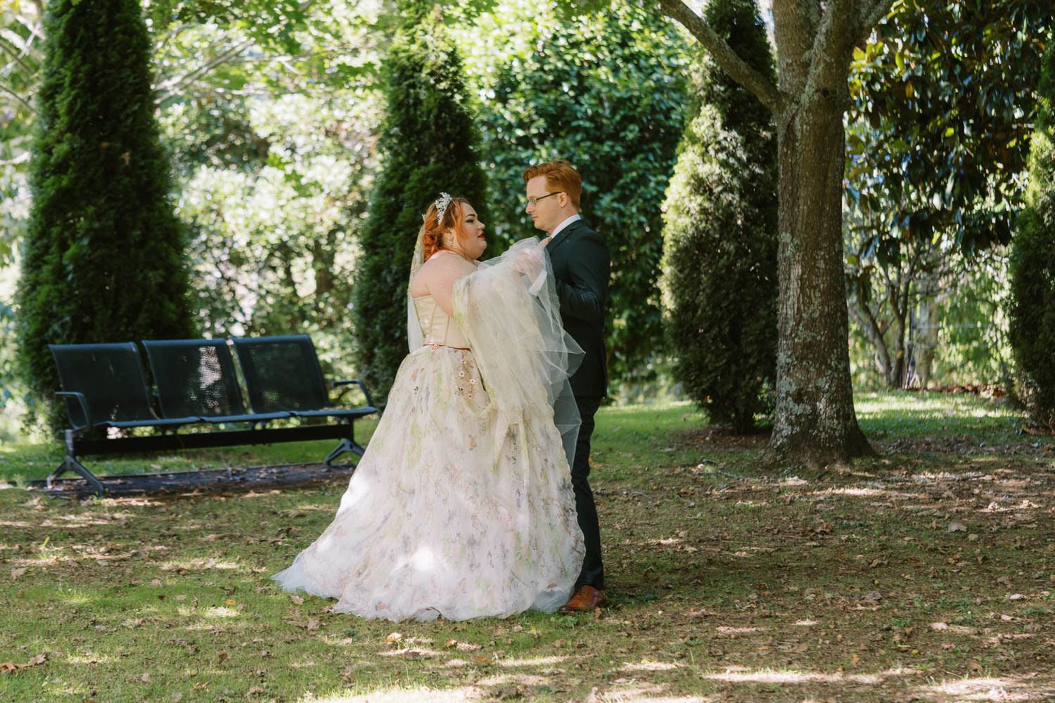 A bride and groom share a romantic moment in a lush green park, with the bride wearing a flowing floral gown.