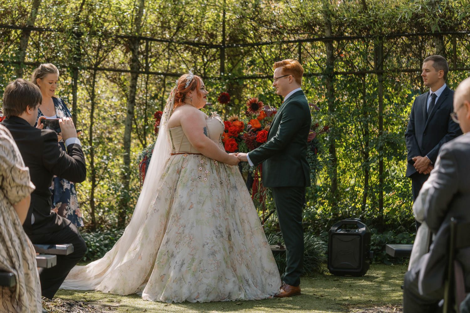 Bride in floral gown and groom in teal suit exchange vows at an outdoor garden wedding ceremony.