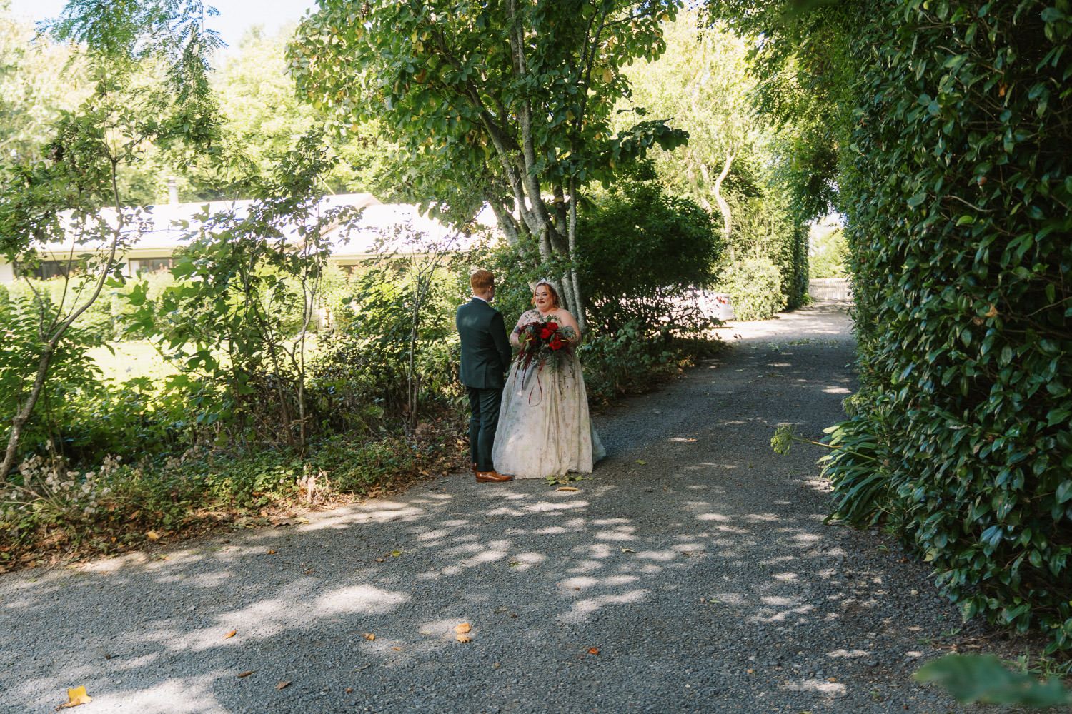 A bride and groom walk hand in hand down a tree-lined gravel path during their outdoor wedding ceremony.