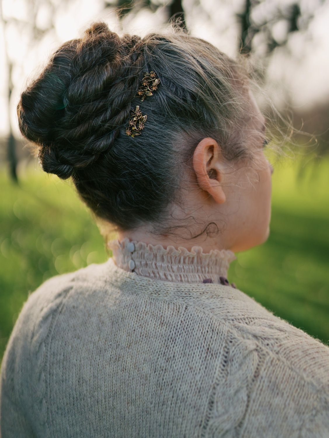 Woman with braided updo hair accessory and neck tattoo wearing cream cable-knit sweater outdoors.