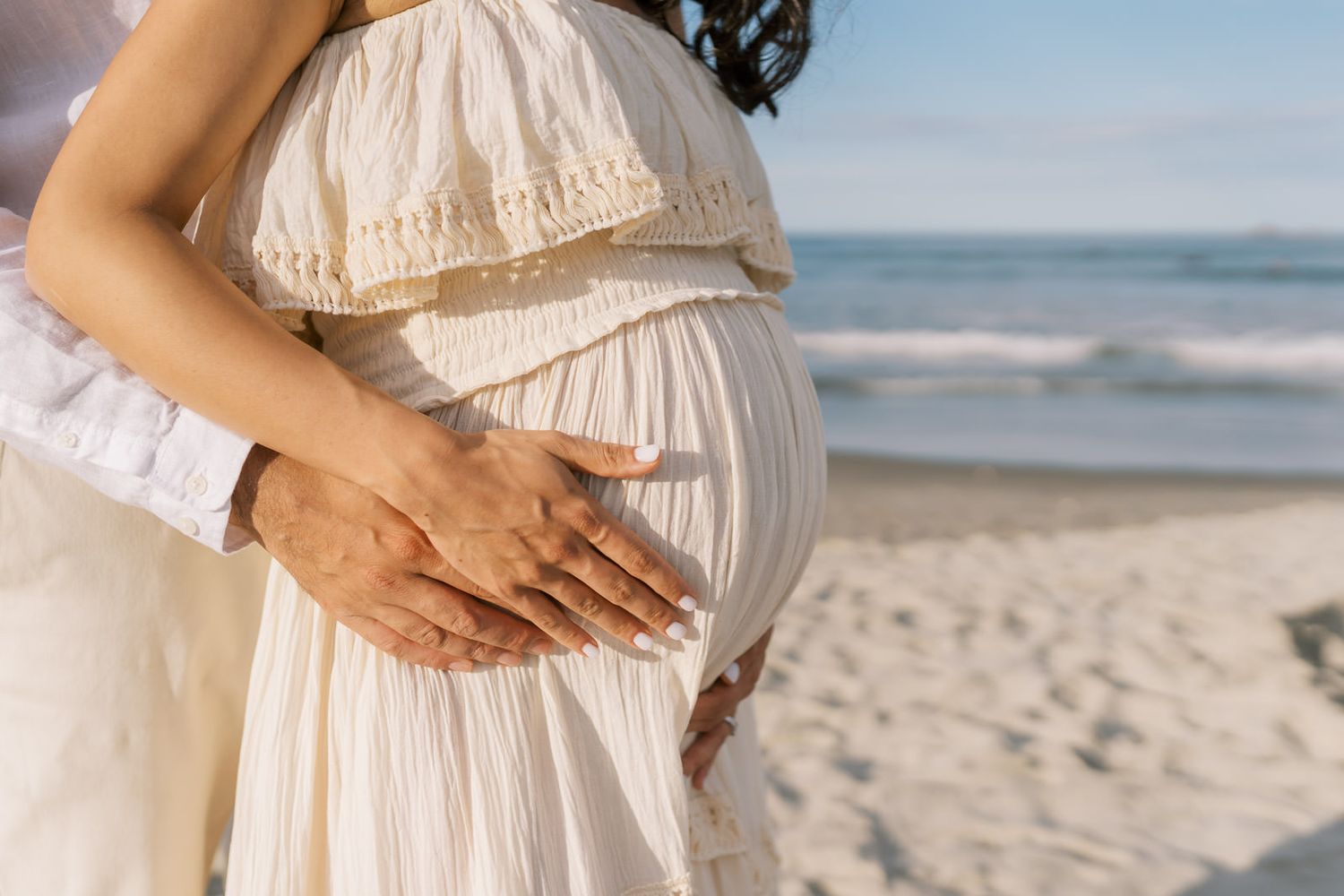 Pregnant woman in white dress at beach with partner's hands on baby bump, ocean waves in background.