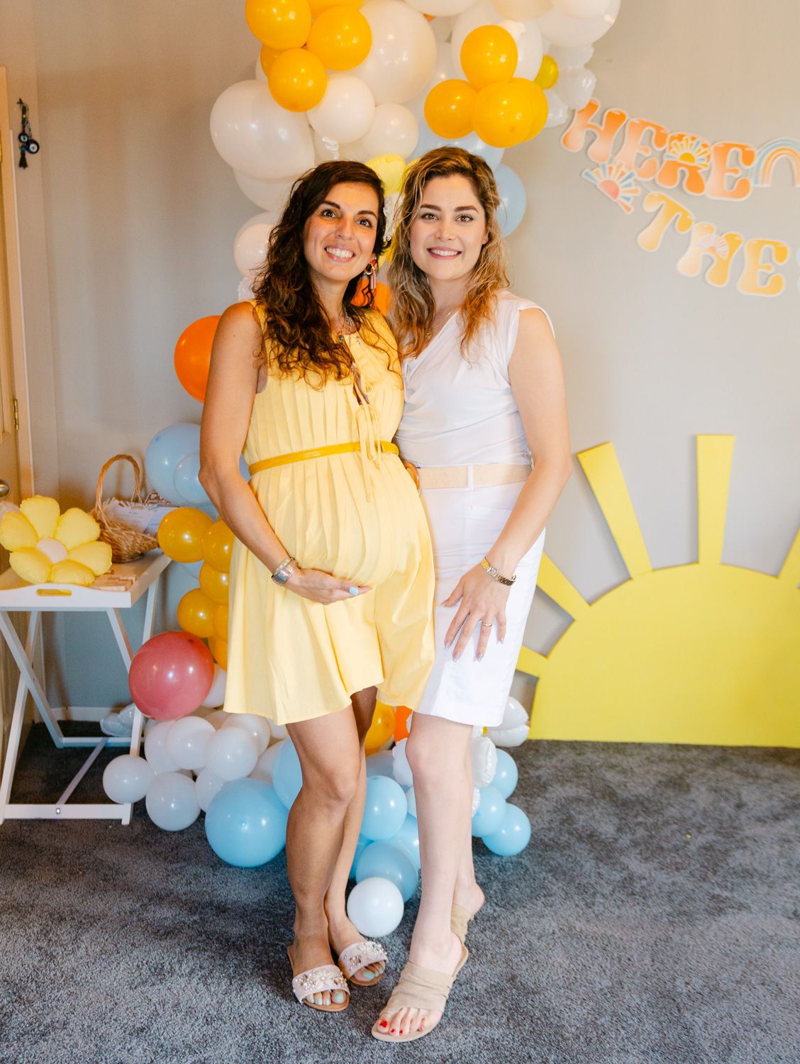 Two women posing at a sunny baby shower with yellow balloon decorations and a sun backdrop.