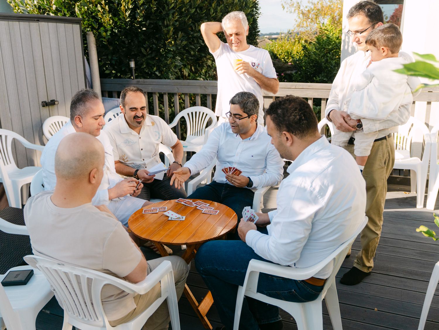Group of men in white shirts playing cards at outdoor table on wooden deck with white plastic chairs.