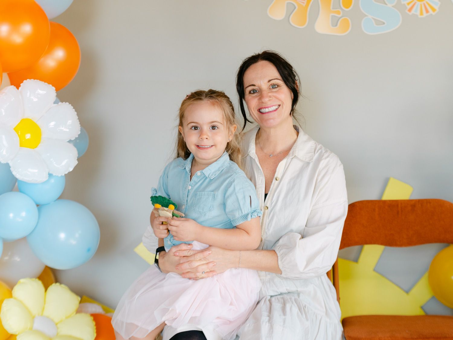 A woman and young girl smile together at a colorful balloon party backdrop with daisy and pastel decorations.