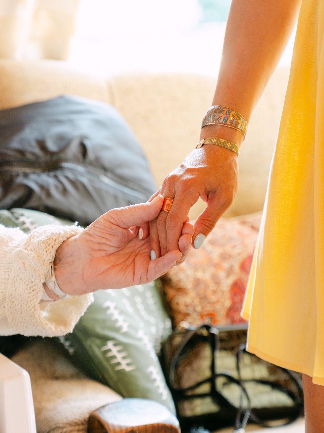 Two women holding hands tenderly, one wearing a gold bracelet and yellow dress, the other in a white sweater with a ring.
