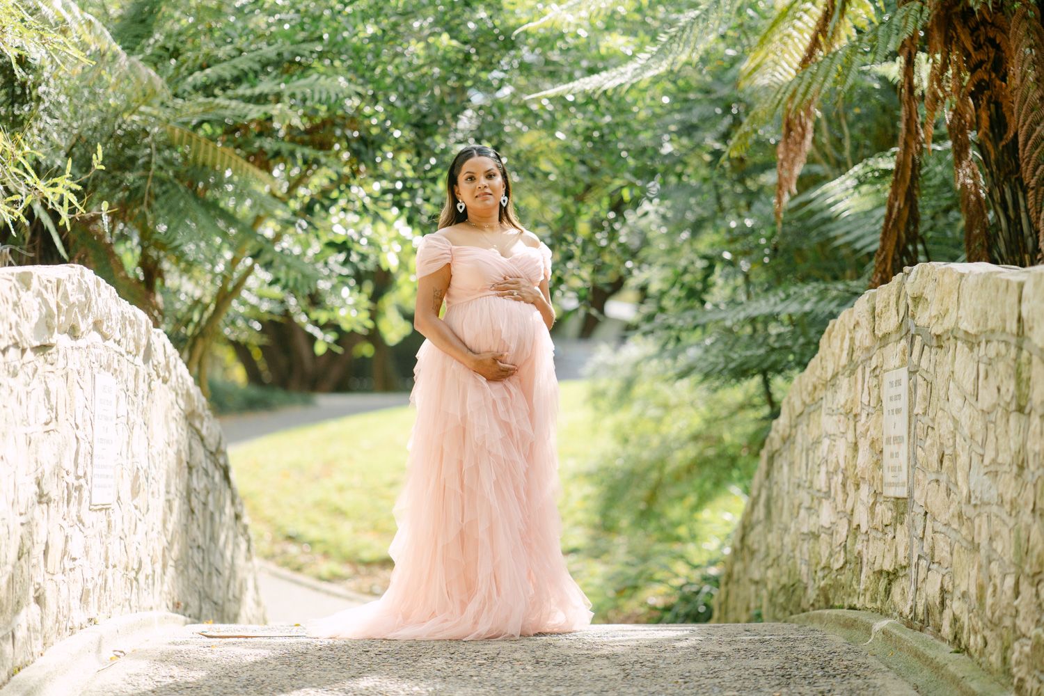 Pregnant woman in pink tulle maternity gown poses on stone bridge surrounded by lush green trees.