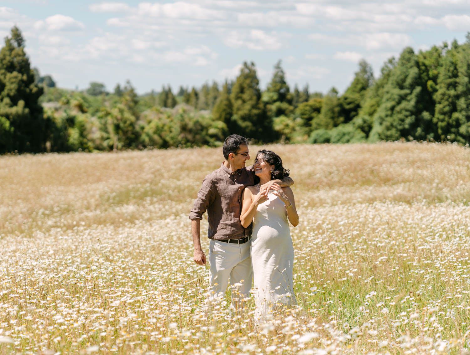 Couple embracing in a sunlit daisy field with lush green trees in the background on a beautiful day.
