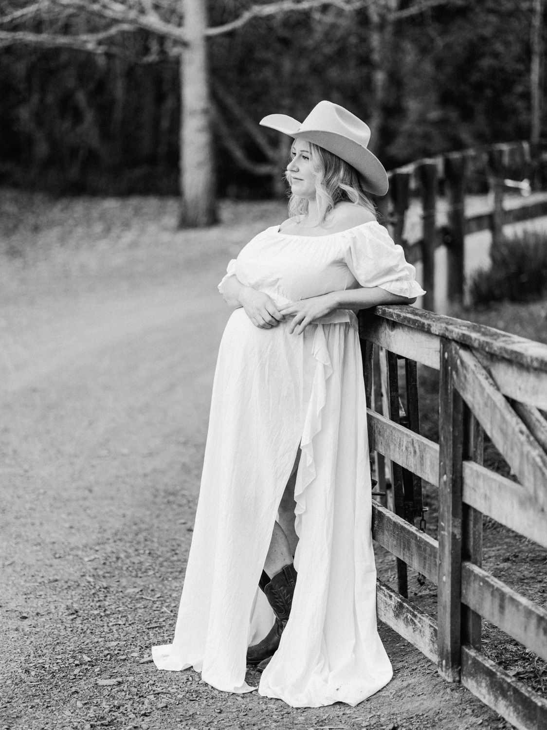 Black and white photo of a woman in a white off-shoulder gown and cowboy hat leaning on a wooden fence outdoors.